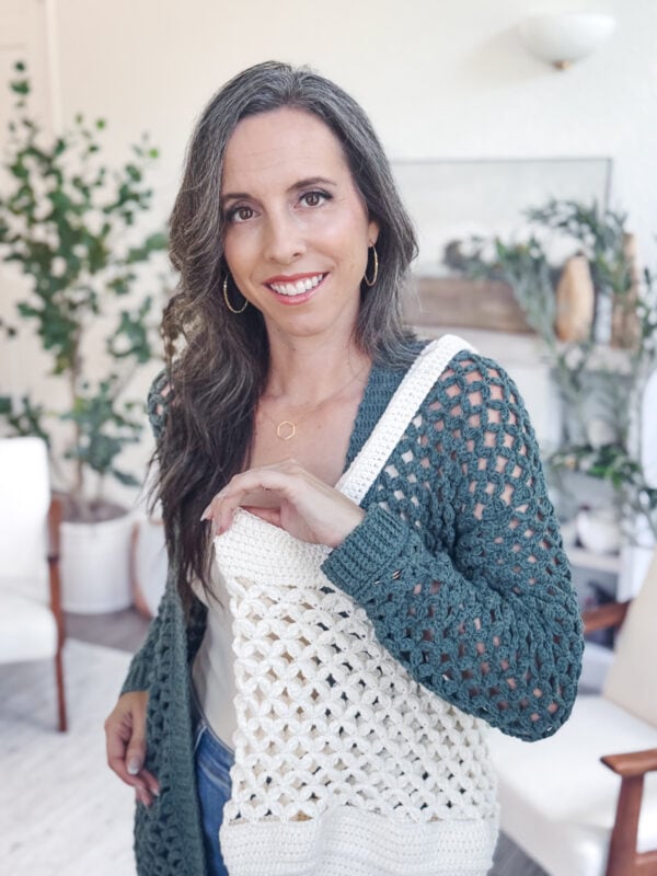 A woman with long brown hair wearing a teal and white Granny Stitch crocheted cardigan stands indoors, smiling at the camera. There are plants and modern decor in the background.
