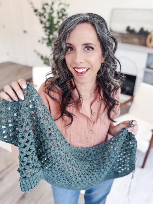 A woman with long wavy gray hair, wearing a peach shirt, holds up a green crocheted garment indoors and smiles at the camera.