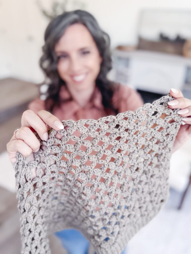 A person holds up a piece of beige crochet fabric, displaying the textured stitch pattern. The softly lit indoor setting highlights their progress as they learn how to crochet the Circle Leaf Stitch.