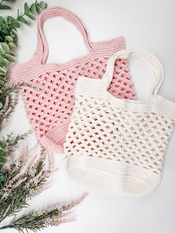 Two crocheted tote bags, one pink and one white, made with a classic Granny Stitch pattern, are laid on a white surface with green and pink foliage arranged nearby.