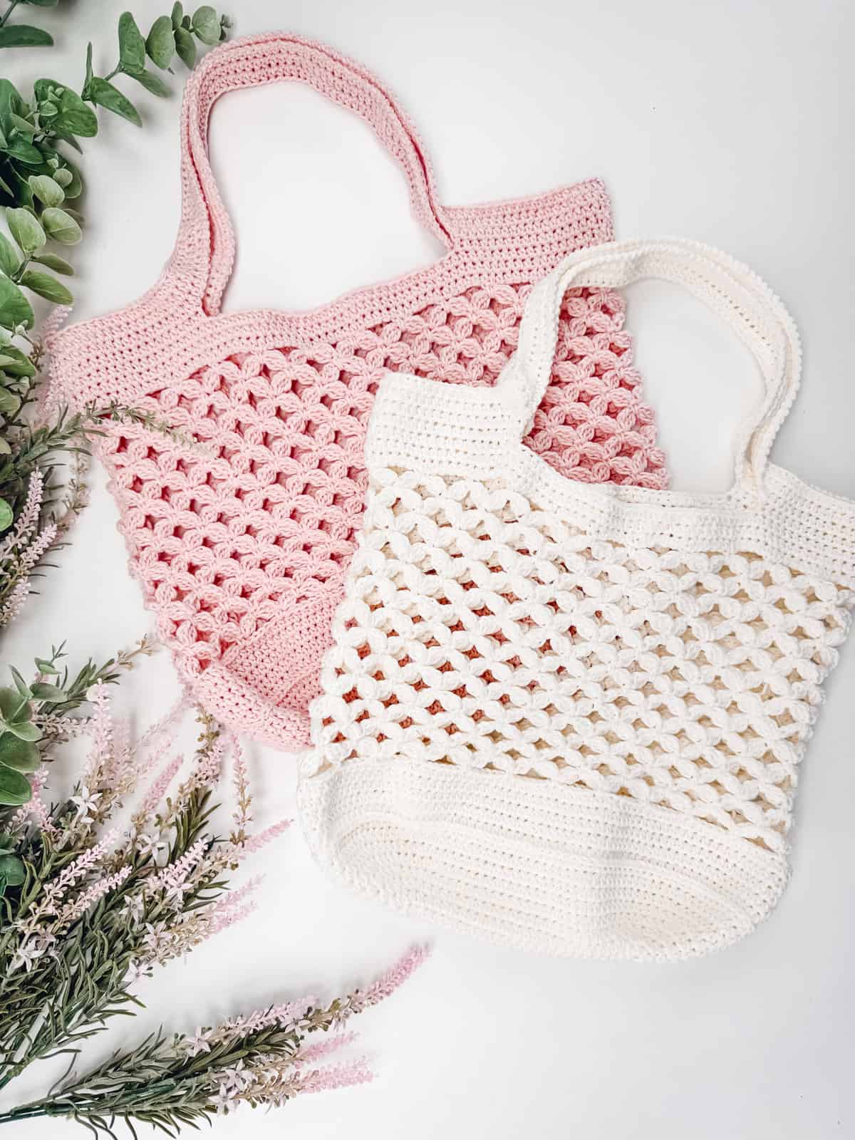 Two crocheted tote bags, one pink and one white, made with a classic Granny Stitch pattern, are laid on a white surface with green and pink foliage arranged nearby.