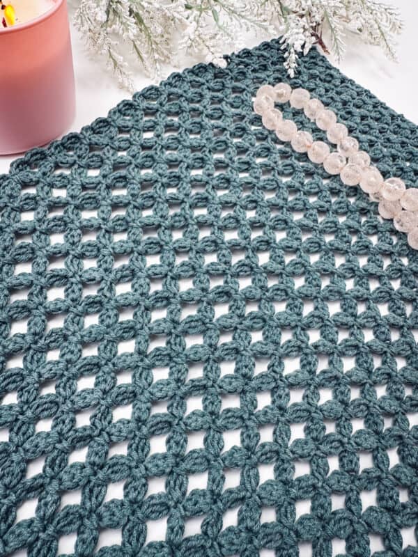Teal openwork crochet fabric displayed on a flat surface with a pink candle, a beaded bracelet, and frosted greenery in the background.