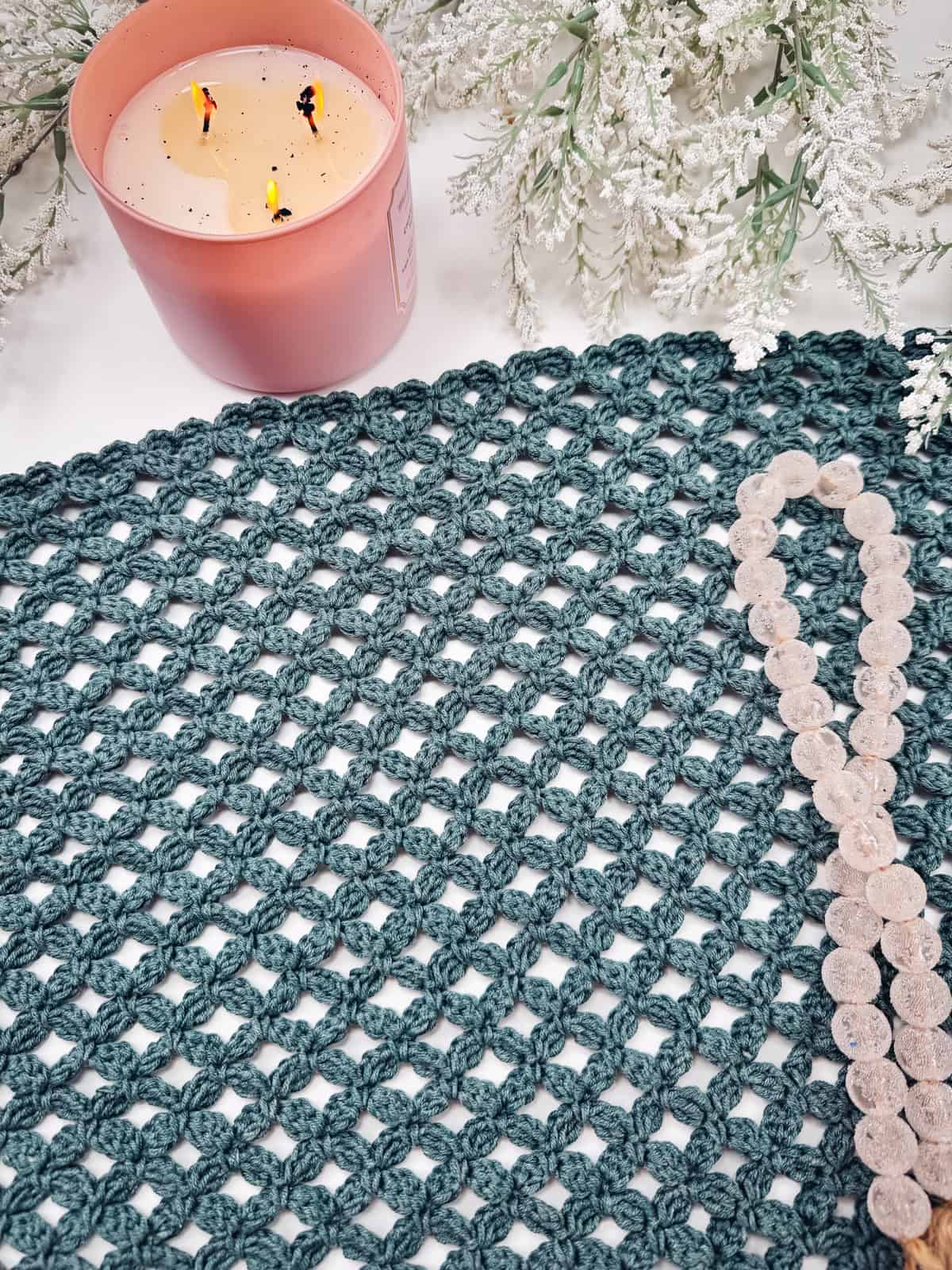 A teal crocheted fabric, showcasing the Circle Leaf Stitch, with a beaded necklace rests beside a pink candle and white frosted foliage on a white surface.