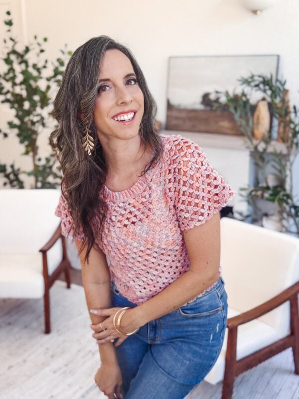 A woman with long brown hair wearing a pink granny stitch crochet top and blue jeans smiles while posing in a bright, modern room with plants and chairs.