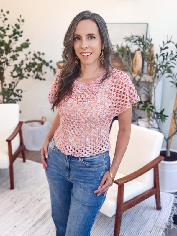 A woman with long brown hair stands indoors, wearing a pink granny stitch crocheted top and blue jeans, with plants and white chairs in the background.