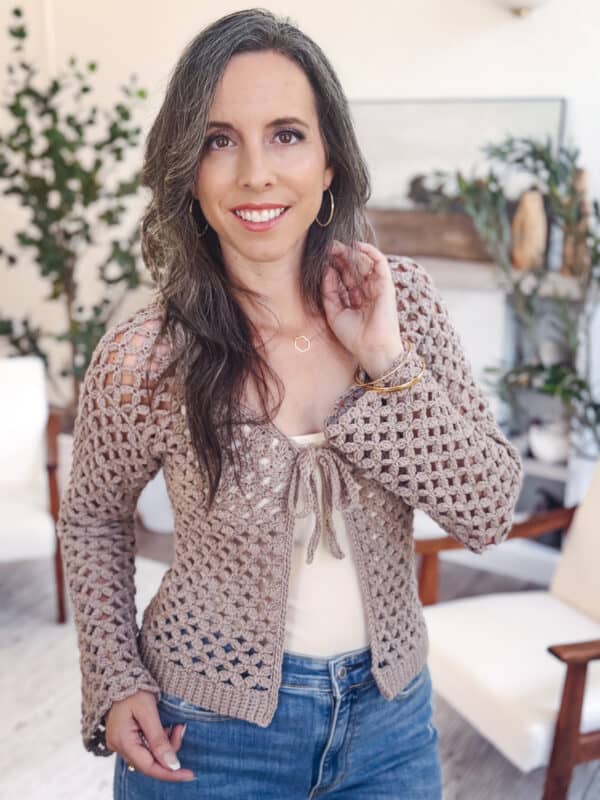 A woman with long brown hair, wearing a Granny Stitch crocheted cardigan over a white top and blue jeans, stands indoors in a room decorated with plants and white chairs.