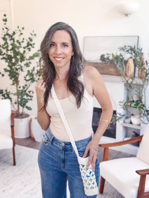 A woman with long, wavy brown hair wearing a beige tank top and blue jeans stands indoors, holding a Free Crochet Water Bottle Holder bag. There are plants and chairs in the background.