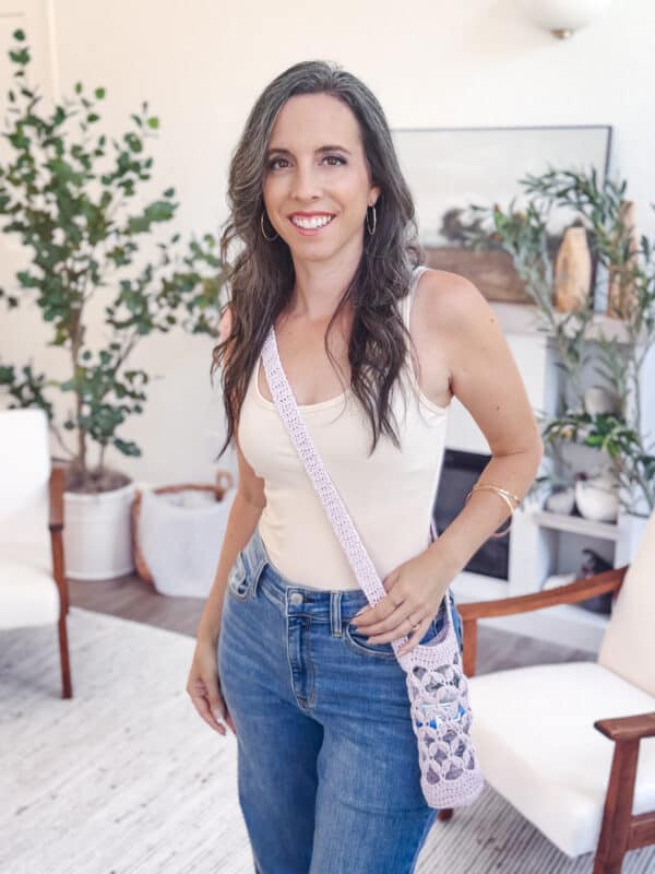 Woman with long brown hair wearing a beige tank top and blue jeans, standing indoors with a crocheted Granny Stitch crossbody bag, smiling at the camera.