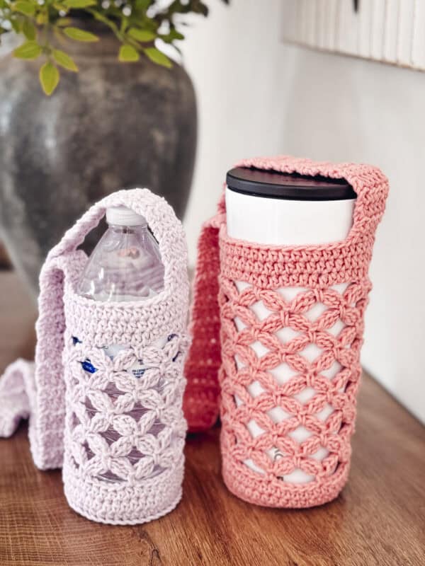 Two reusable bottles, one plastic and one metal, are placed in crocheted holders with shoulder straps; the holders are pink and lavender and rest on a wooden surface.