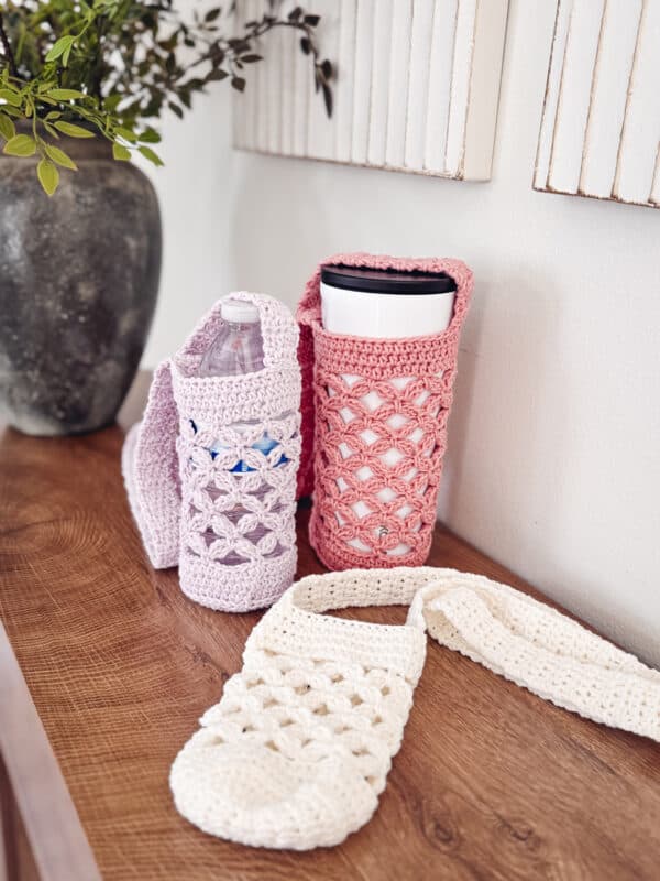 Three crochet water bottle holders in pink, lavender, and white sit on a wooden surface; two contain bottles, one is empty. A vase with green foliage is in the background.
