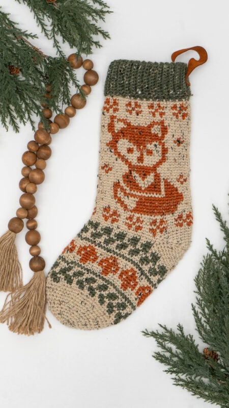 Knitted Christmas stocking with a fox pattern, green top cuff, and decorative wooden bead garland, surrounded by evergreen branches on a white background.