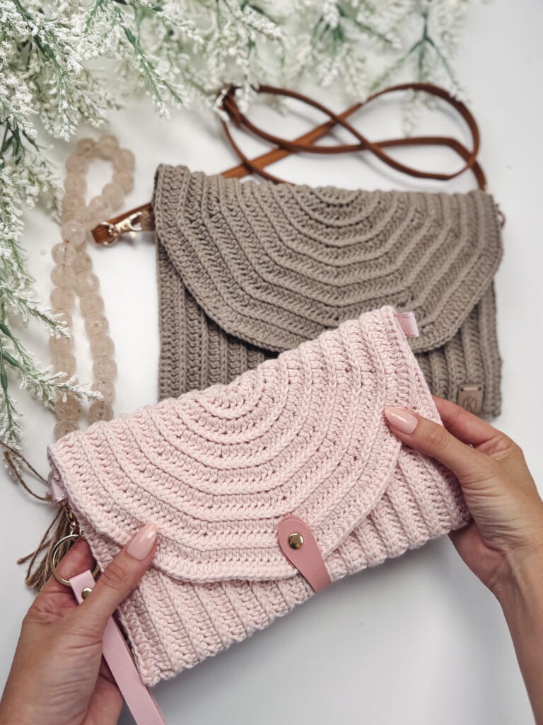 Two crocheted handbags, made using an easy crochet clutch pattern, are displayed on a white surface with green foliage. The pink, beginner-friendly envelope bag is held in someone's hands.