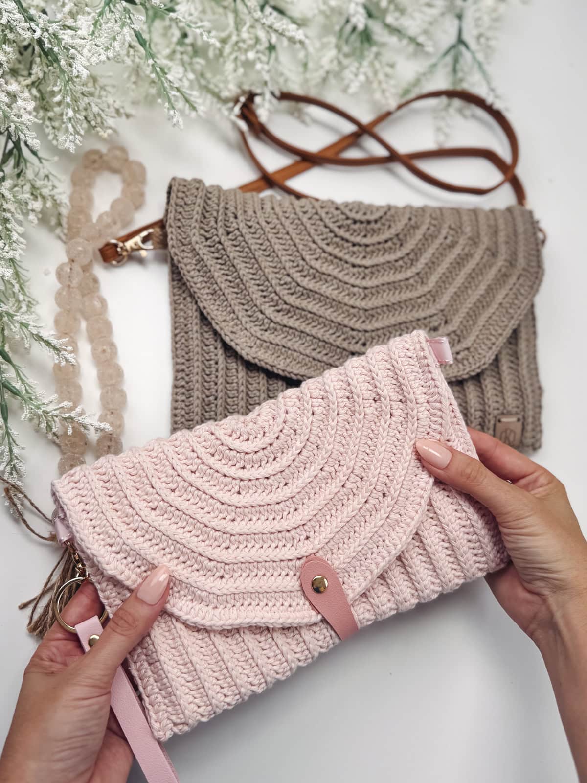 Two crocheted handbags, made using an easy crochet clutch pattern, are displayed on a white surface with green foliage. The pink, beginner-friendly envelope bag is held in someone's hands.