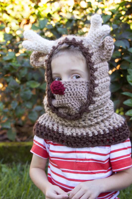 A child wearing a red-striped shirt and a knitted reindeer hat with antlers and a red nose stands outdoors in front of greenery.