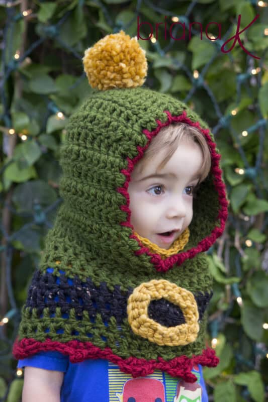 A child wears a green crocheted hooded cape with red and yellow trim, a yellow pom-pom on top, and a decorative buckle, standing in front of greenery with string lights.