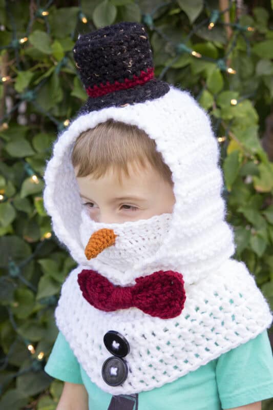 A young child wears a crocheted snowman costume hood with a carrot nose, red bow tie, black buttons, and a black hat, standing in front of greenery with string lights.