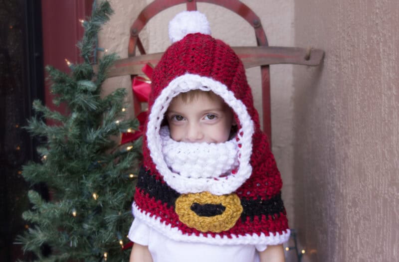 A young child wearing a crocheted Santa Claus hooded cape stands in front of a small decorated Christmas tree and a wooden chair.