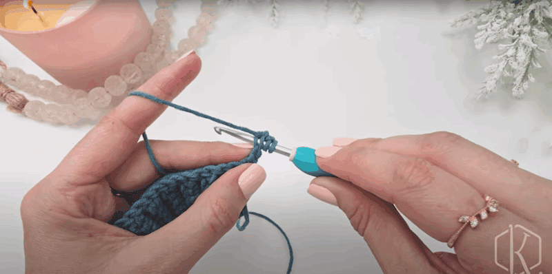 A person uses a blue-handled crochet hook to work with teal yarn, possibly learning how to crochet the Circle Leaf Stitch. A lit candle, beads, and white decorations are visible in the background.