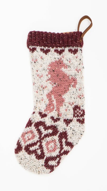 Knitted Christmas stocking with a maroon, pink, and cream pattern, featuring a reindeer design and a brown hanging loop at the top.