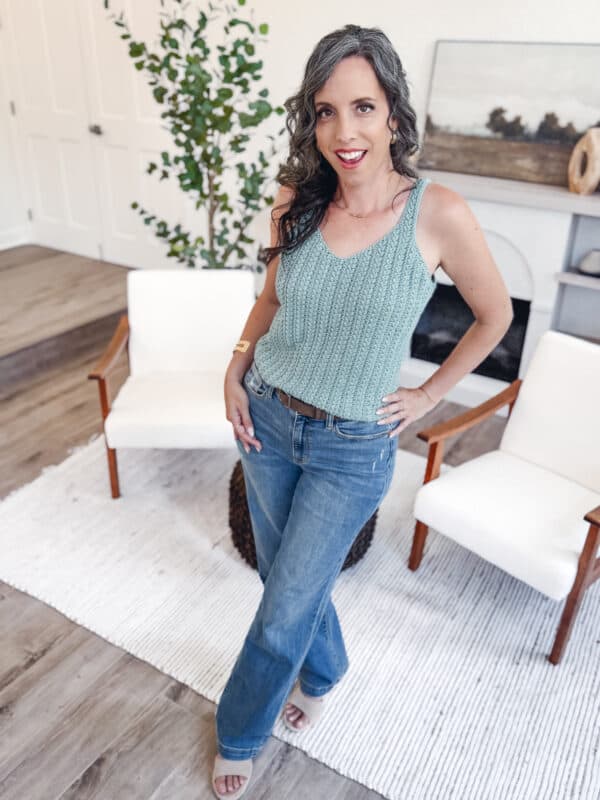 A woman with long, wavy gray hair wearing a teal Wheat Stitch Tank Top - Free Crochet Pattern and blue jeans stands on a white rug in a modern living room.