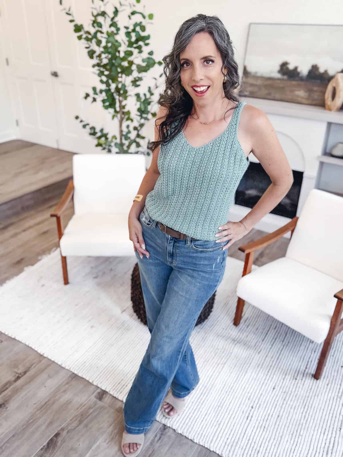 A woman with long, wavy gray hair wearing a teal Wheat Stitch Tank Top - Free Crochet Pattern and blue jeans stands on a white rug in a modern living room.