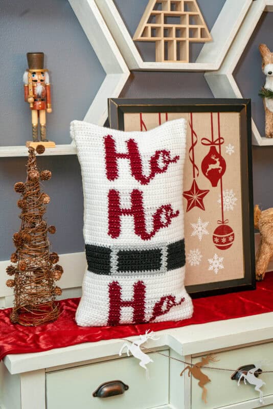 A crocheted pillow with "Ho Ho Ho" and a black belt design sits on a decorated shelf with holiday ornaments, a framed reindeer print, and a nutcracker.