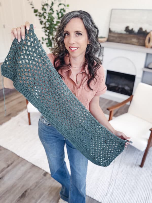 A woman in a pink shirt and jeans holds up a green crocheted rectangular piece indoors, standing on a rug near a white chair and a fireplace.