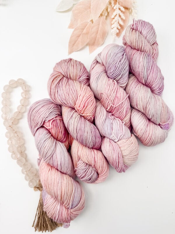 Four skeins of pink and lavender yarn are arranged on a white surface with blush-colored leaves and a beaded tassel nearby.