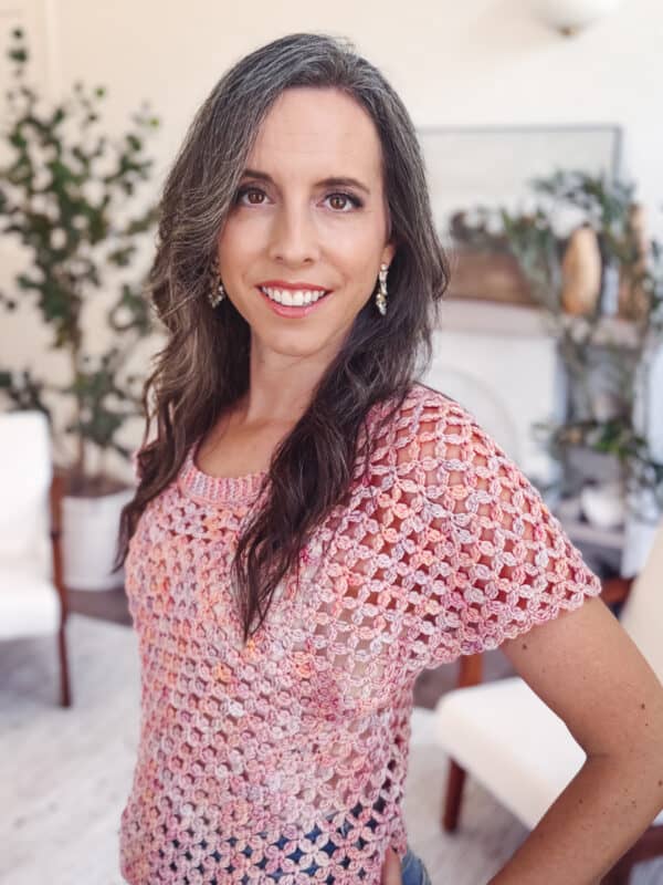 A woman with long brown hair and a pink crocheted top stands indoors, smiling at the camera. There are green plants and white chairs in the background.