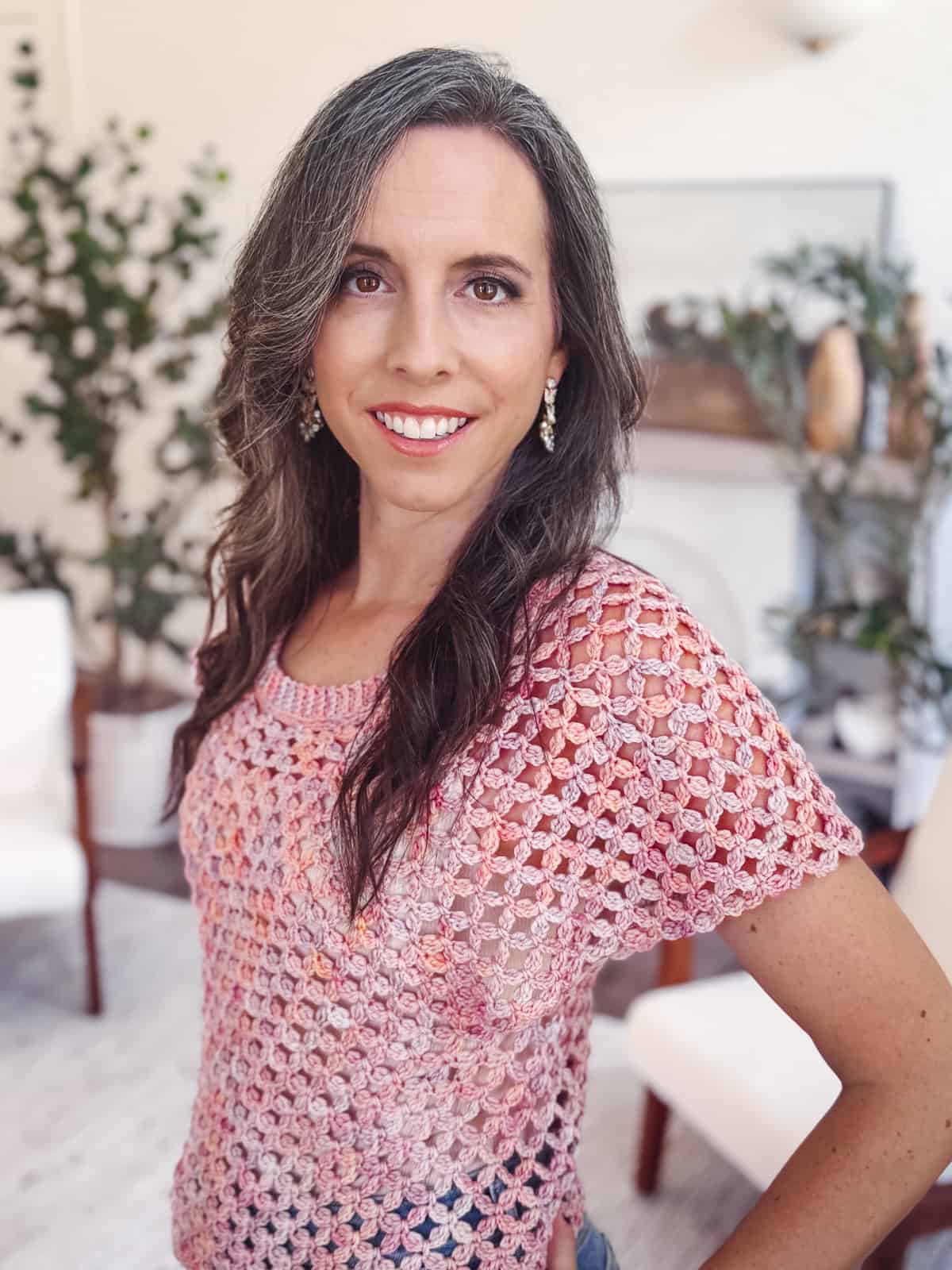 A woman with long brown hair and a pink crocheted top stands indoors, smiling at the camera. There are green plants and white chairs in the background.