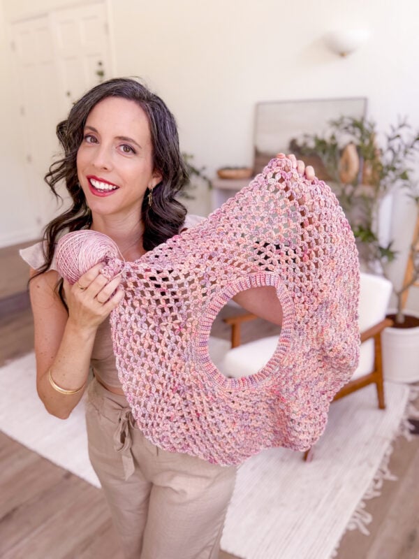Woman holding a crochet project made with pink yarn, smiling at the camera in a well-lit living room.