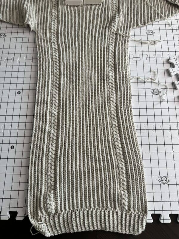 A knitted garment featuring vertical ribbing and cable patterns is laid flat on a blocking mat with a visible grid and blocking pins, demonstrating how blocking crochet projects ensures a professional finish.