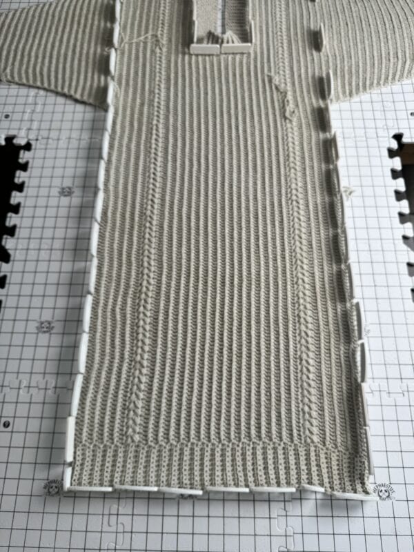 A knitted garment is laid flat on a blocking mat with grid lines; white blocking rods secure the edges, ensuring crochet projects achieve a professional finish.