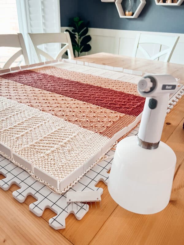 A knitted fabric piece is blocking on foam mats on a table, with a handheld steamer and small brush nearby—perfect tools for blocking crochet projects to achieve a professional finish.