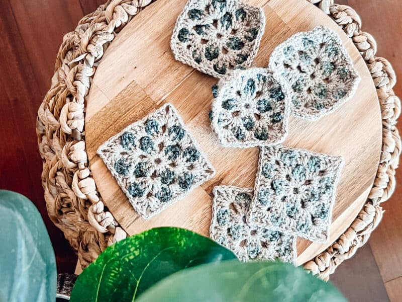 Six crocheted granny squares in muted colors are arranged on a round wooden board with woven edge accents, next to some green leaves in the foreground.