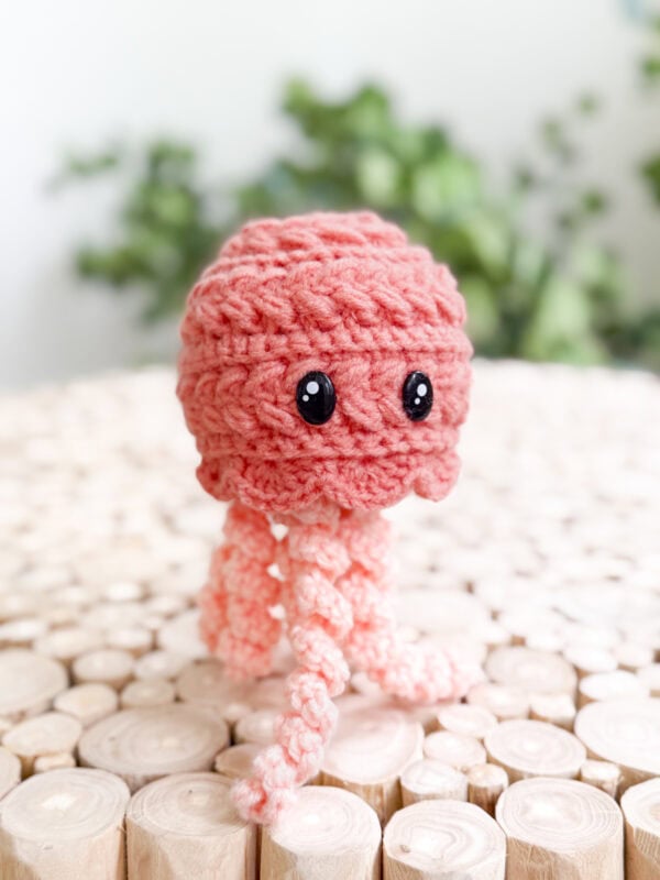 A small, pink crocheted jellyfish with black eyes sits on a wooden surface, with green plants blurred in the background.