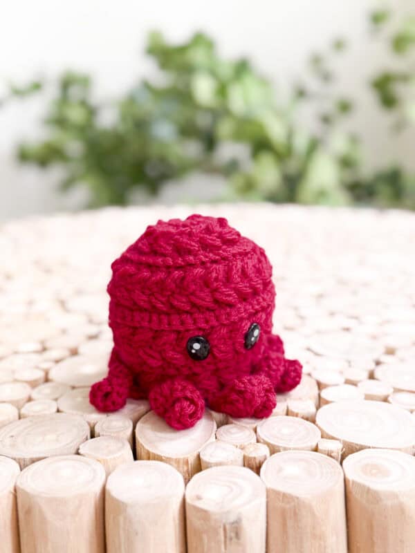 A small, red crocheted octopus with black button eyes sits on a surface made of light wooden logs, with green plants in the background.