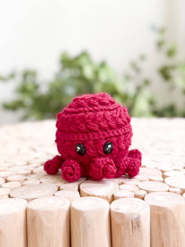 A small red crocheted octopus with black eyes sits on round wooden logs, with blurred green plants in the background—made using a free octopus amigurumi crochet pattern.