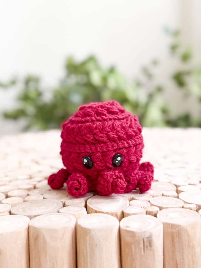 A small red crocheted octopus with black eyes sits on round wooden logs, with blurred green plants in the background—made using a free octopus amigurumi crochet pattern.
