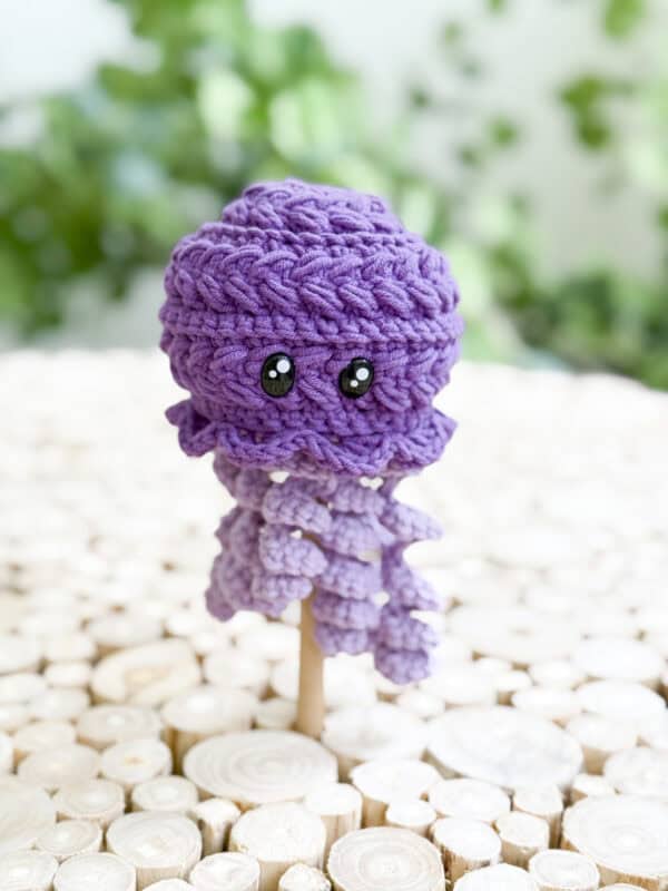 A crocheted purple octopus doll with black eyes is displayed on a wooden stick, set against a light wooden surface and a leafy green background—perfect inspiration for those seeking a free crochet jellyfish amigurumi pattern.