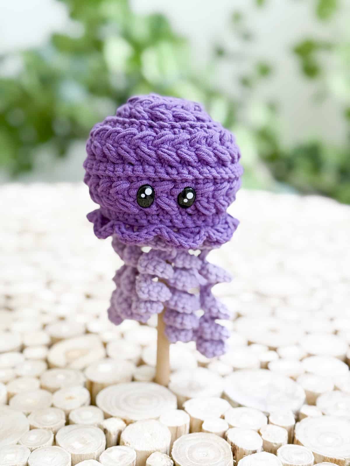 A crocheted purple octopus doll with black eyes is displayed on a wooden stick, set against a light wooden surface and a leafy green background—perfect inspiration for those seeking a free crochet jellyfish amigurumi pattern.