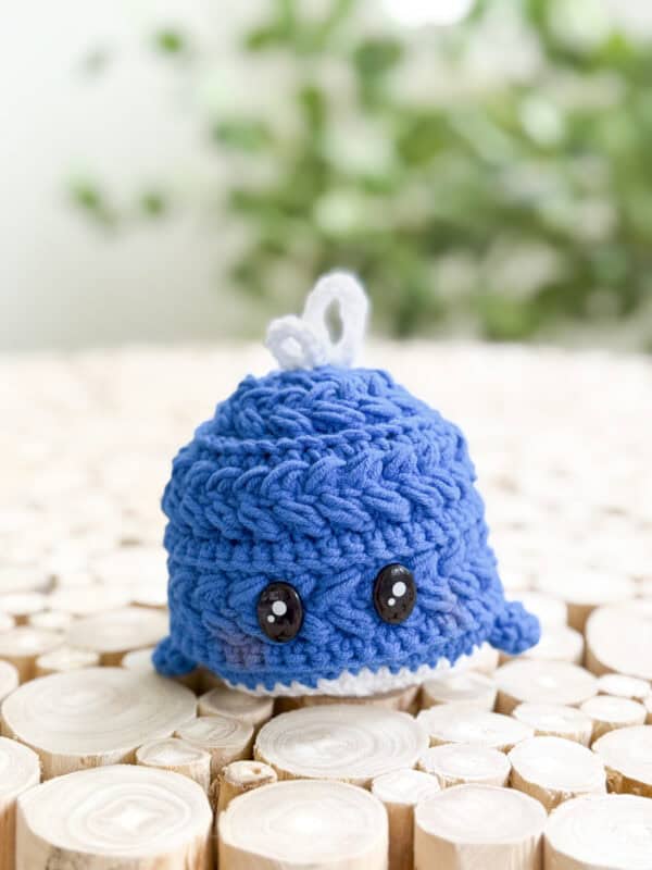 A small blue crocheted whale with black button eyes, made using the Free Wheat Stitch Crochet Whale Pattern, sits on a surface of round wooden slices against a blurred green background.
