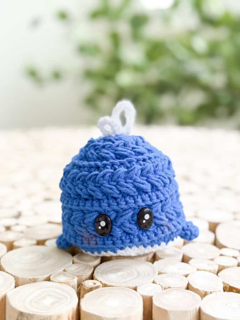 A small blue crocheted whale with black button eyes, made using the Free Wheat Stitch Crochet Whale Pattern, sits on a surface of round wooden slices against a blurred green background.