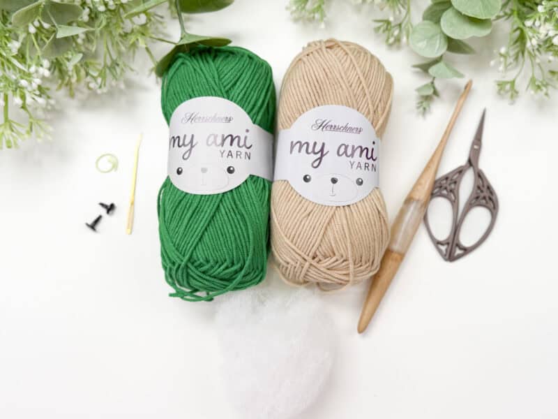 Two skeins of green and beige yarn, crochet hook, scissors, stuffing, and other crochet tools are arranged on a white surface with greenery in the background.