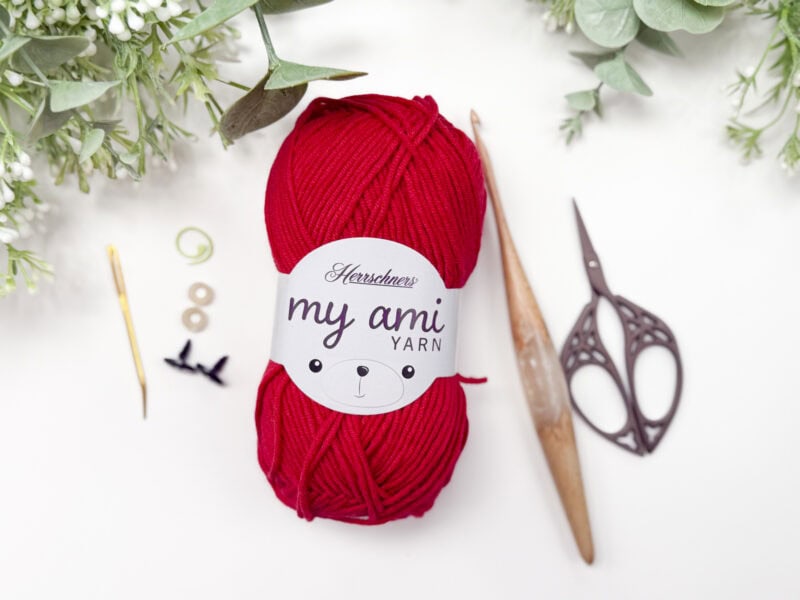 A ball of red "my ami YARN" with scissors, a crochet hook, needles, stitch markers, and green foliage arranged on a white background.