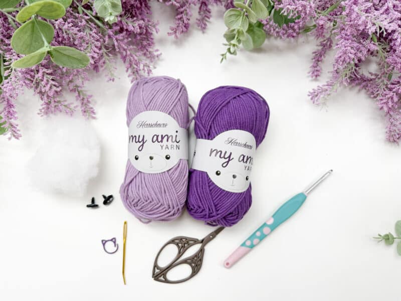 Two skeins of yarn, one light purple and one dark purple, are displayed with a crochet hook, scissors, stuffing, safety eyes, a yarn needle, and stitch marker on a white surface.