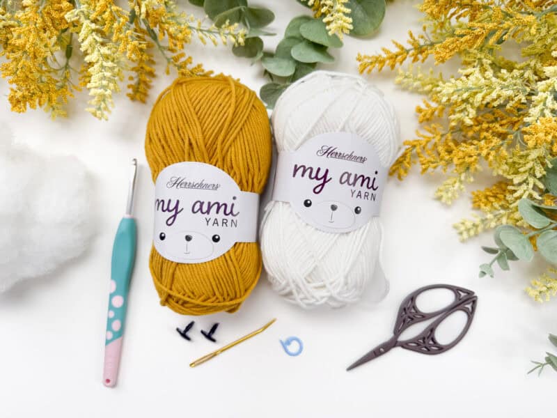 Two skeins of yarn, crochet hook, needle, small scissors, stitch marker, safety eyes, and fiberfill, arranged on a white surface with yellow flowers and green leaves around.