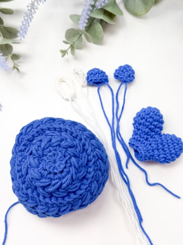 Blue crochet pieces and a heart shape, along with white and blue yarn-tipped strands, are arranged on a white surface with green and lavender foliage in the background.