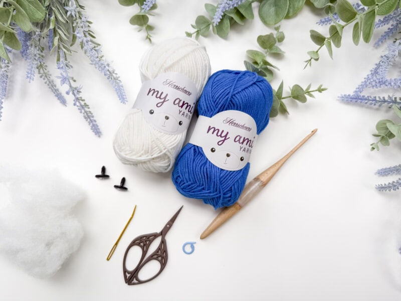 Two skeins of yarn, crochet hook, scissors, stuffing, safety eyes, and other small tools arranged on a white surface surrounded by greenery and lavender flowers.