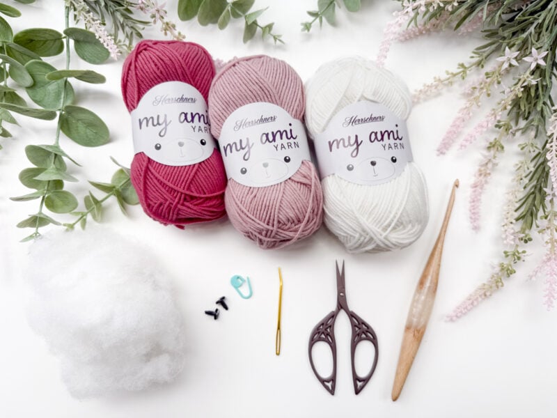 Three skeins of yarn, fiberfill, safety eyes, stitch marker, yarn needle, scissors, and a crochet hook are arranged on a white surface with green and pink foliage.
